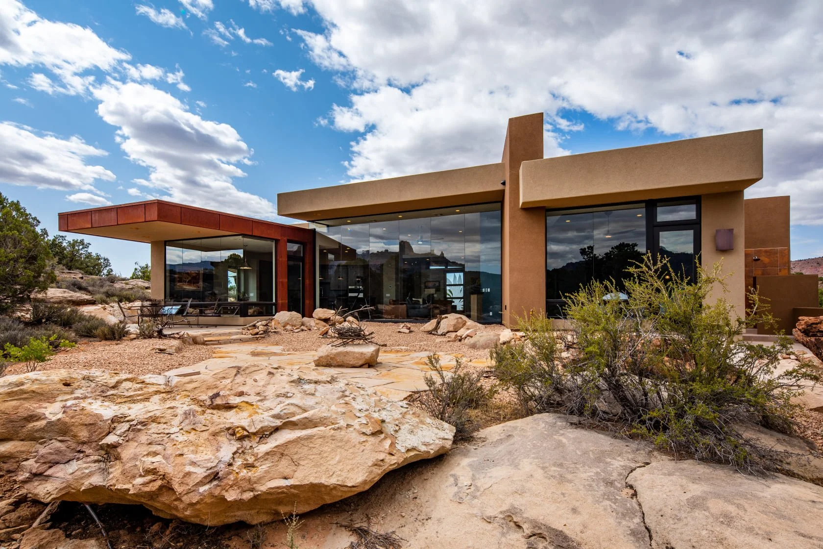 Modern house with large glass windows in a desert landscape with rocks and sparse vegetation under a partly cloudy sky.