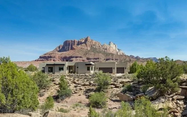 Modern house in a desert landscape with a mountain in the background, green shrubs, and a bright blue sky.
