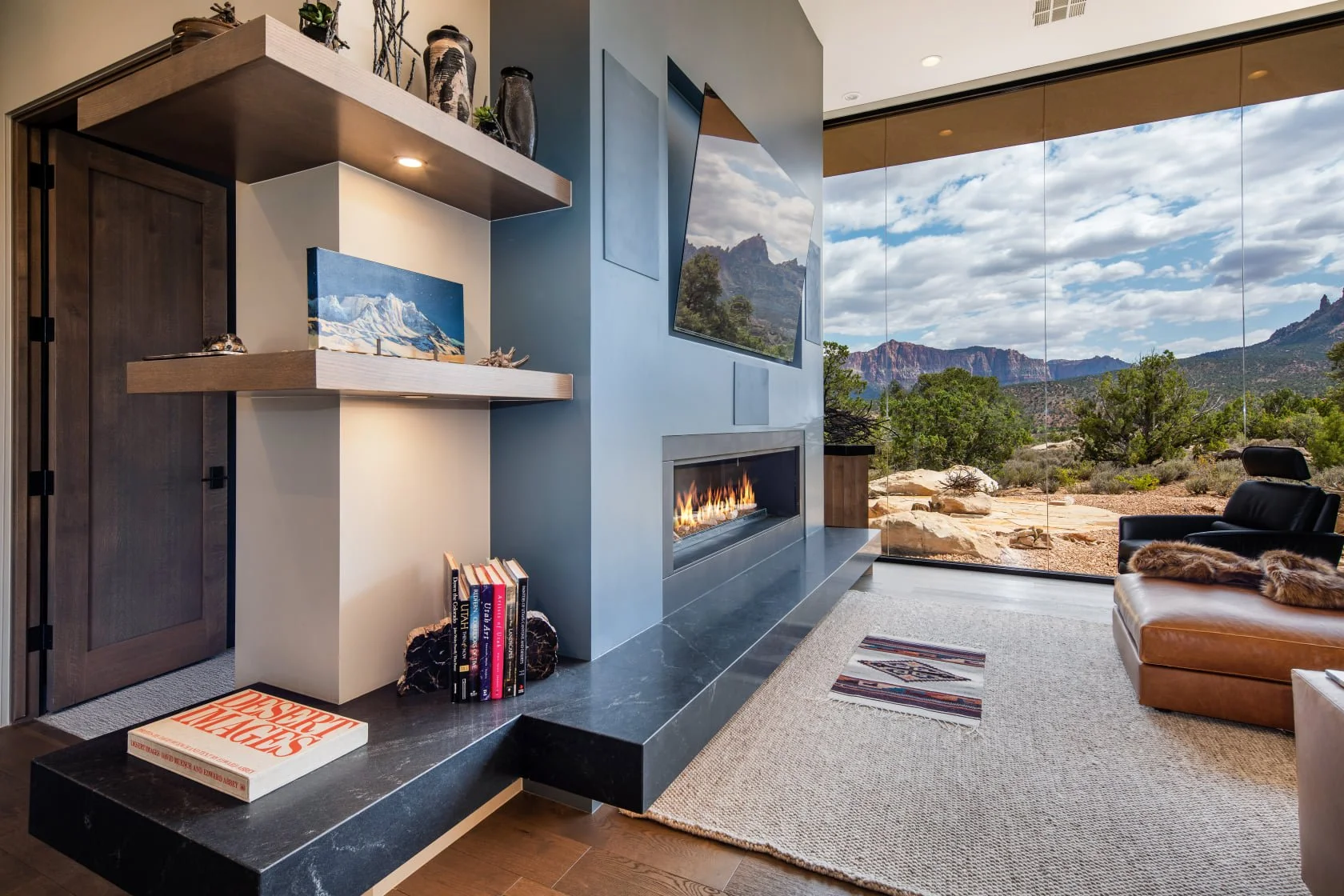 Modern living room with a mountain view through large glass windows, a mounted flat-screen TV, a fireplace, and a tan leather couch.