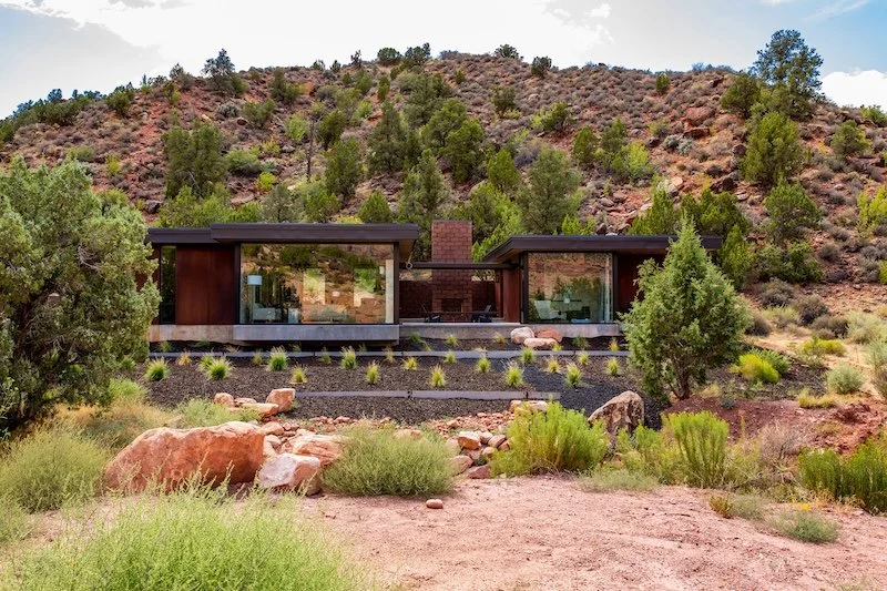 A modern house with large glass windows and a flat roof, situated in a desert landscape with hills and sparse trees.
