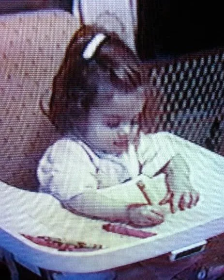 Young girl sitting in a high chair, drawing on paper with a pencil.