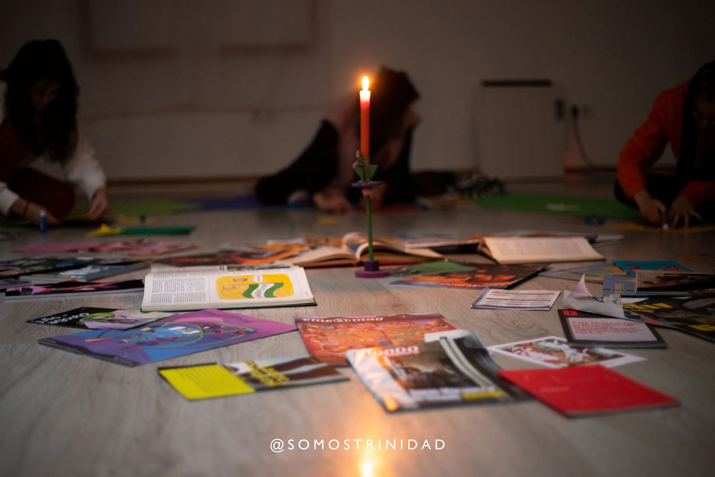 Personas sentadas en el suelo rodeadas de revistas, libros y papeles, con una vela encendida en centro de la mesa, en un ambiente oscuro.