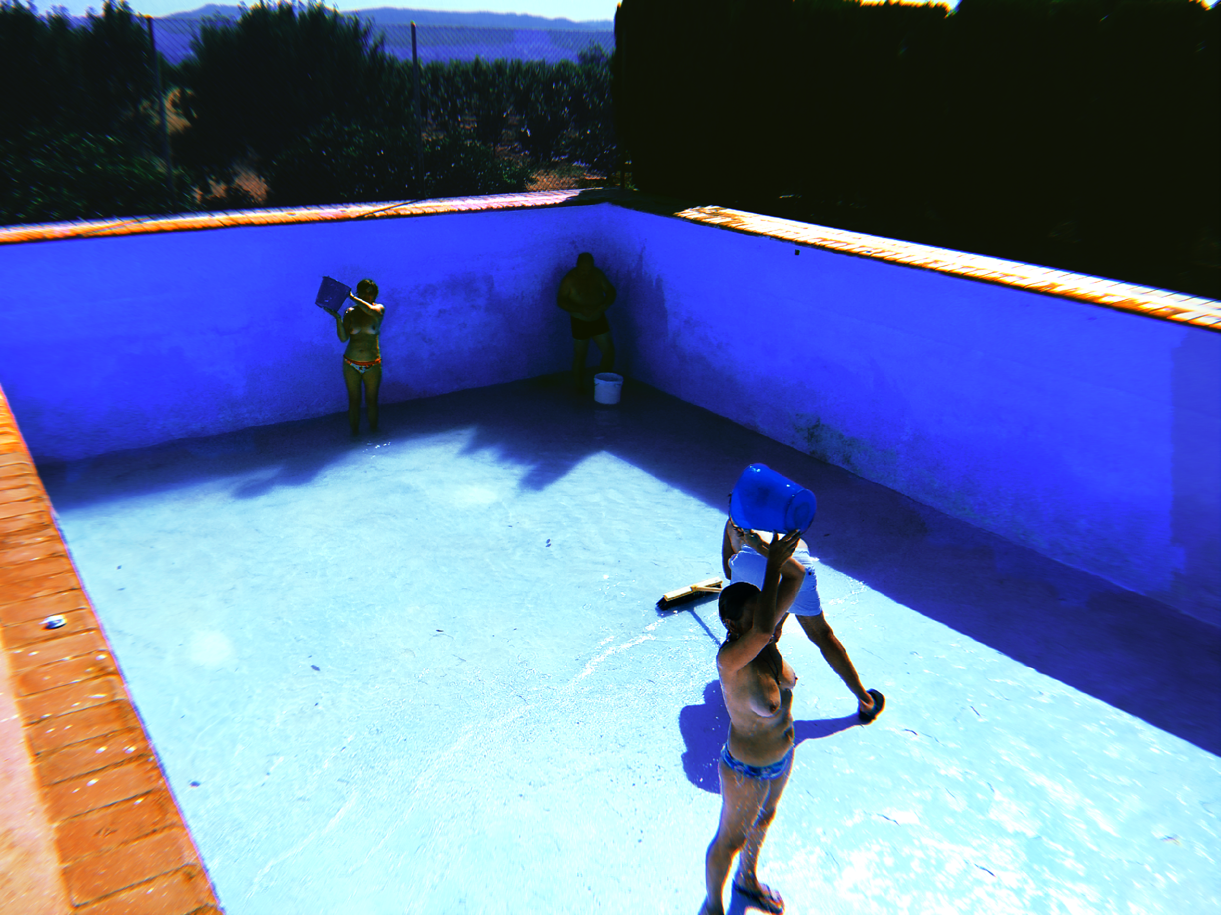 Personas en una alberca sin agua limpiándola, con ropa de baño y herramientas de limpieza, en un entorno rural con árboles y montañas al fondo.