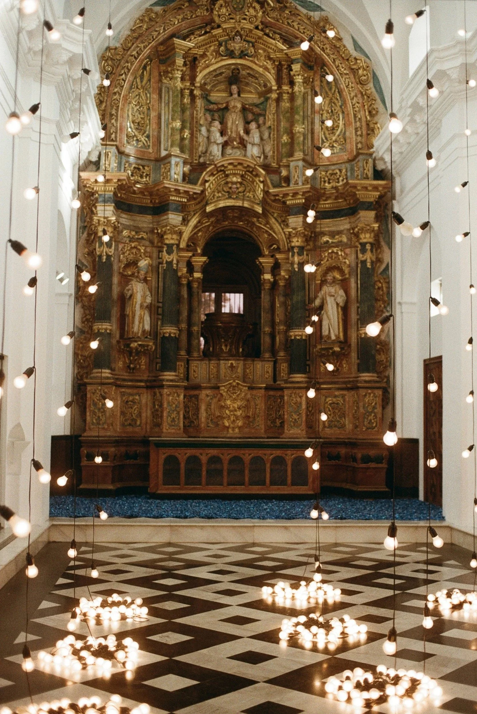 Interiores de una iglesia con altar dorado y luces decorativas suspendidas con focos en el techo y en el suelo.