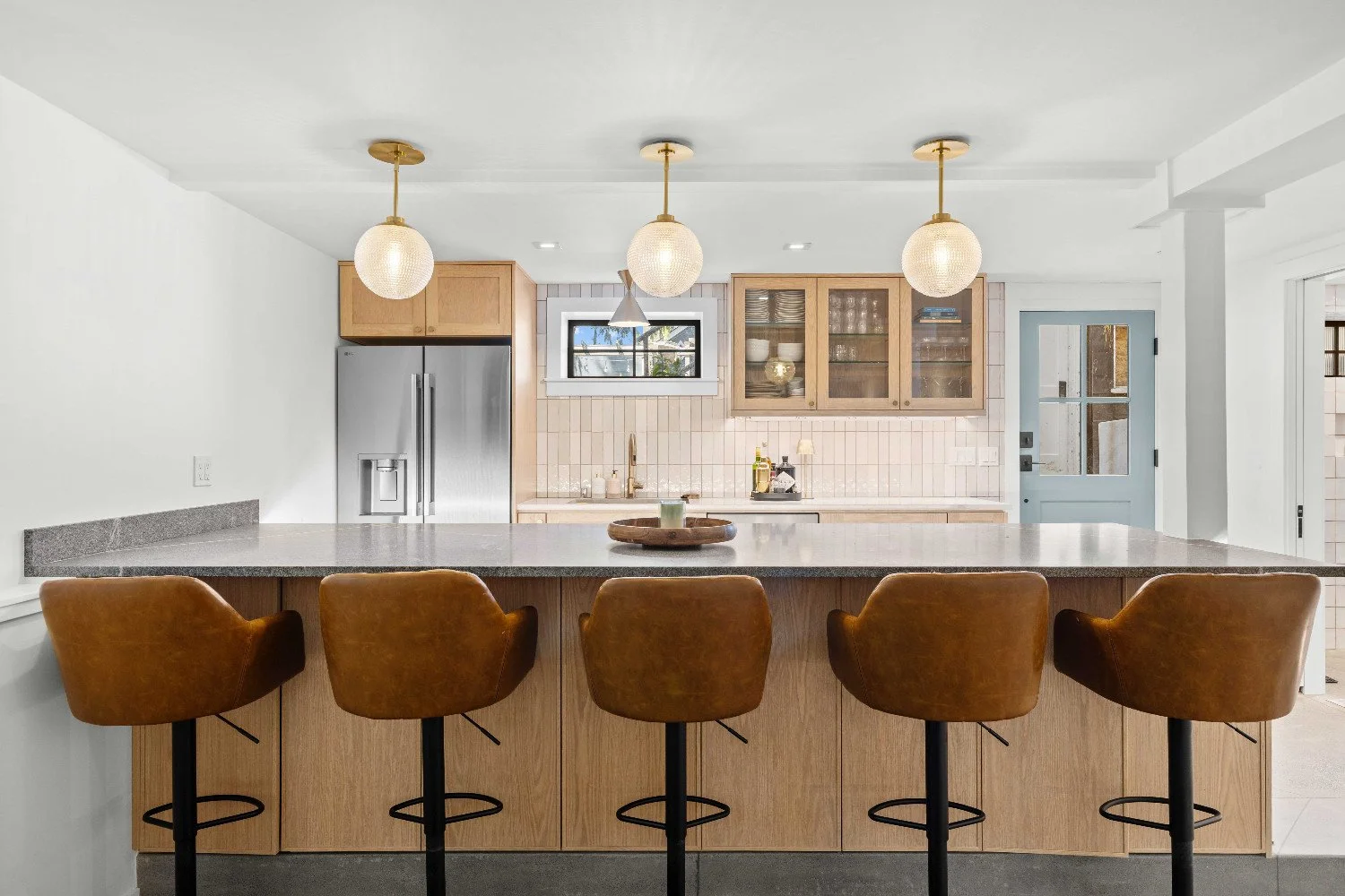 Modern kitchen with a large central island, brown leather barstools, light wood cabinetry, stainless steel refrigerator, white tile backsplash, small window, and decorative pendant lights.