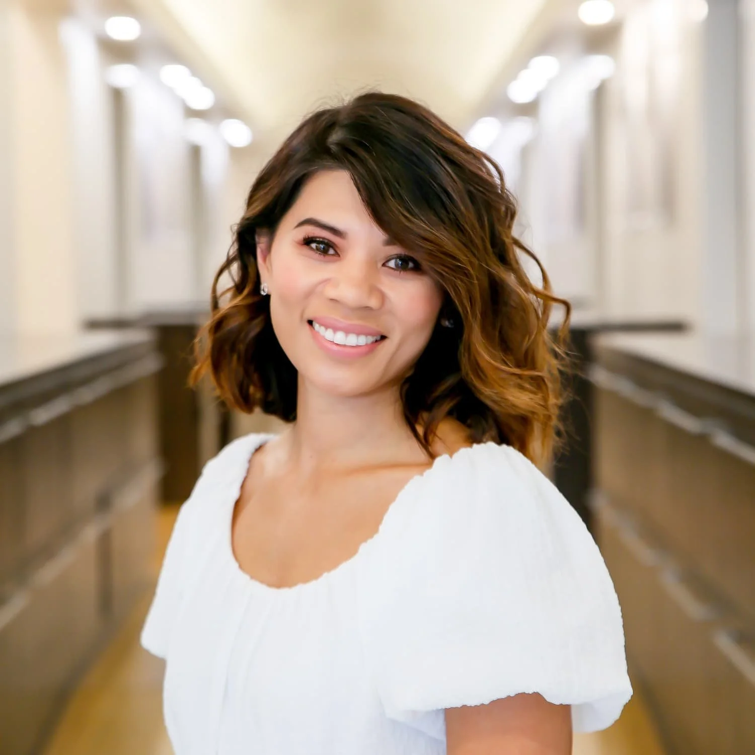 Smiling woman with shoulder-length wavy hair, wearing a white blouse, standing in a brightly lit hallway.