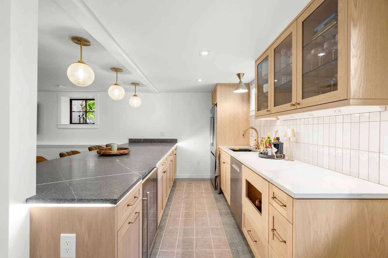 Modern kitchen with light wood cabinets, white countertops, and a tiled backsplash, featuring hanging pendant lights and a small window.