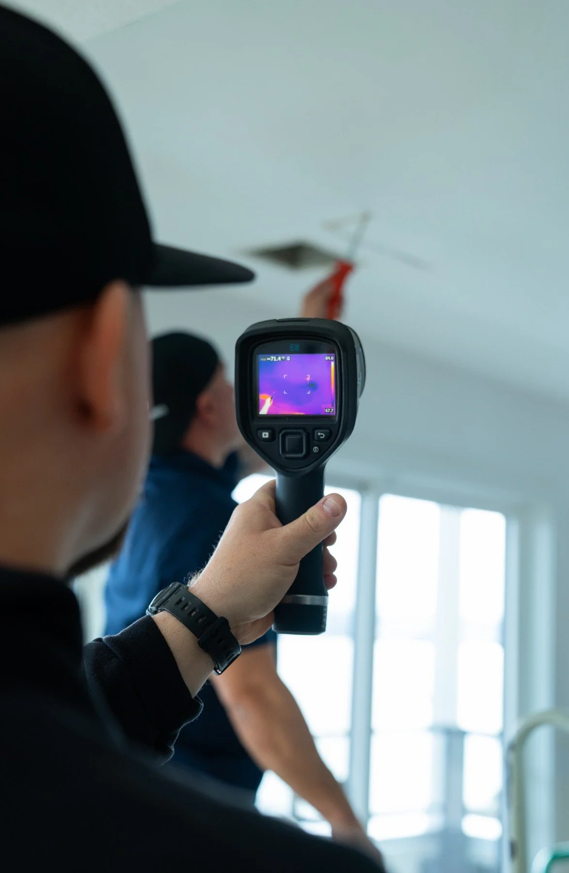 Person in black cap using a thermal imaging camera to inspect a ceiling vent, with another person in the background.