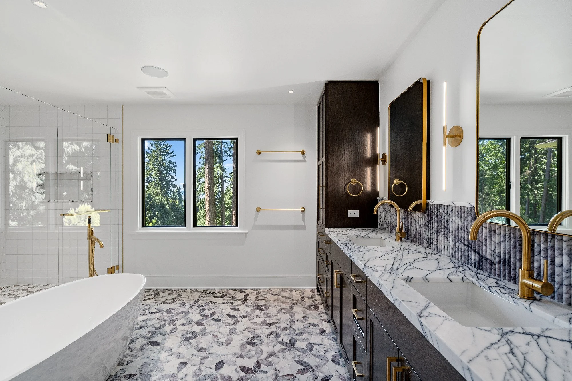Modern bathroom with double marble vanity, gold fixtures, dark wooden cabinets, large windows, a freestanding bathtub, and a walk-in shower with glass enclosure.