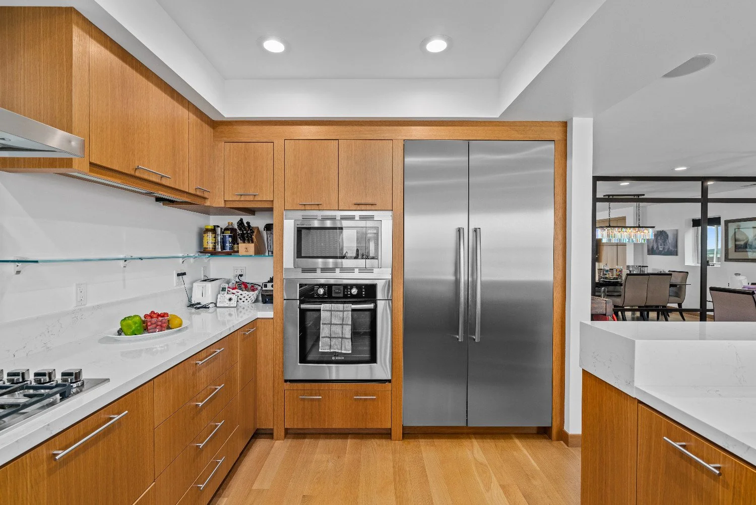 Modern kitchen with wooden cabinets, stainless steel refrigerator, oven, microwave, white countertops, and a glass shelf with kitchen supplies. Part of an open dining and living area is visible in the background.