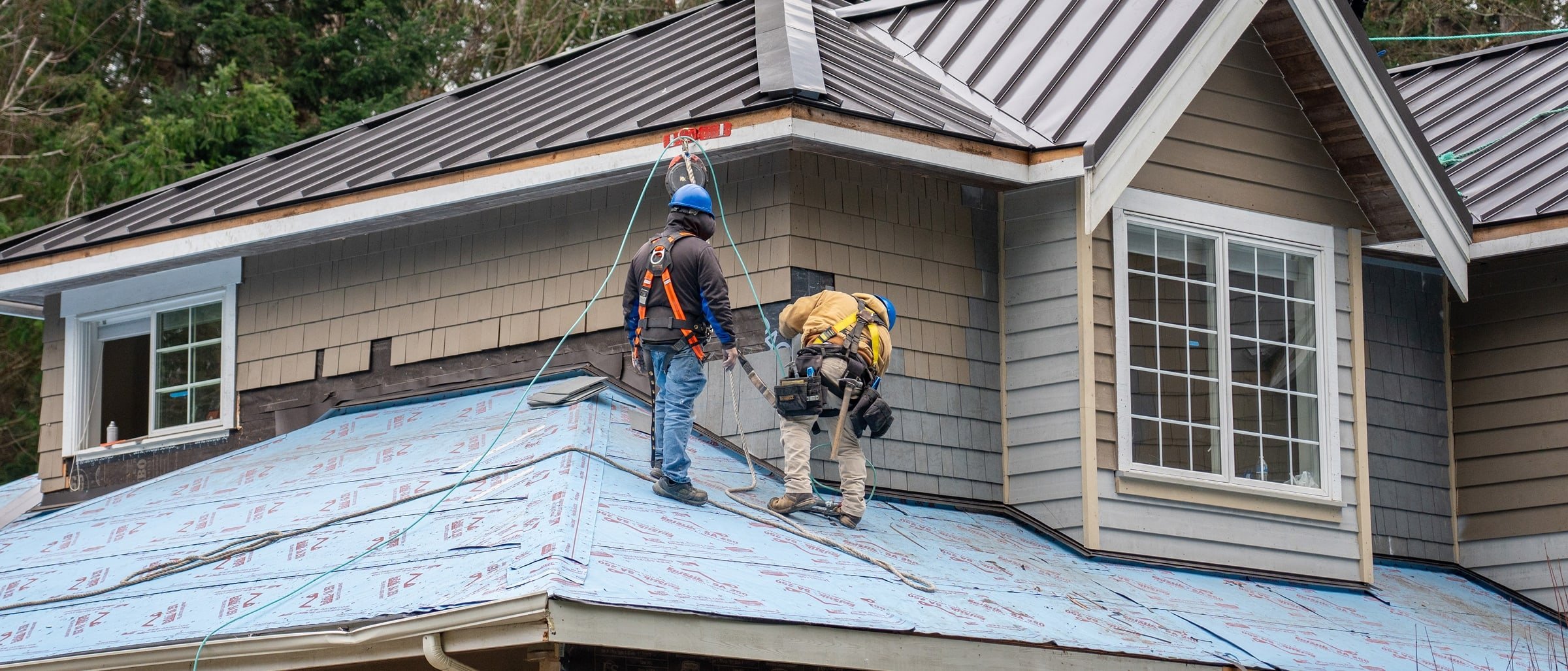 Two construction workers on a sloped roof, wearing safety harnesses and helmets, working on the installation of roofing material on a house with beige and gray siding and white window frames.