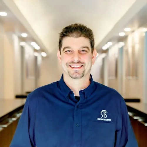 Smiling man with dark hair and beard, wearing a blue button-down shirt with company logo, standing in hallway with lights on ceiling.