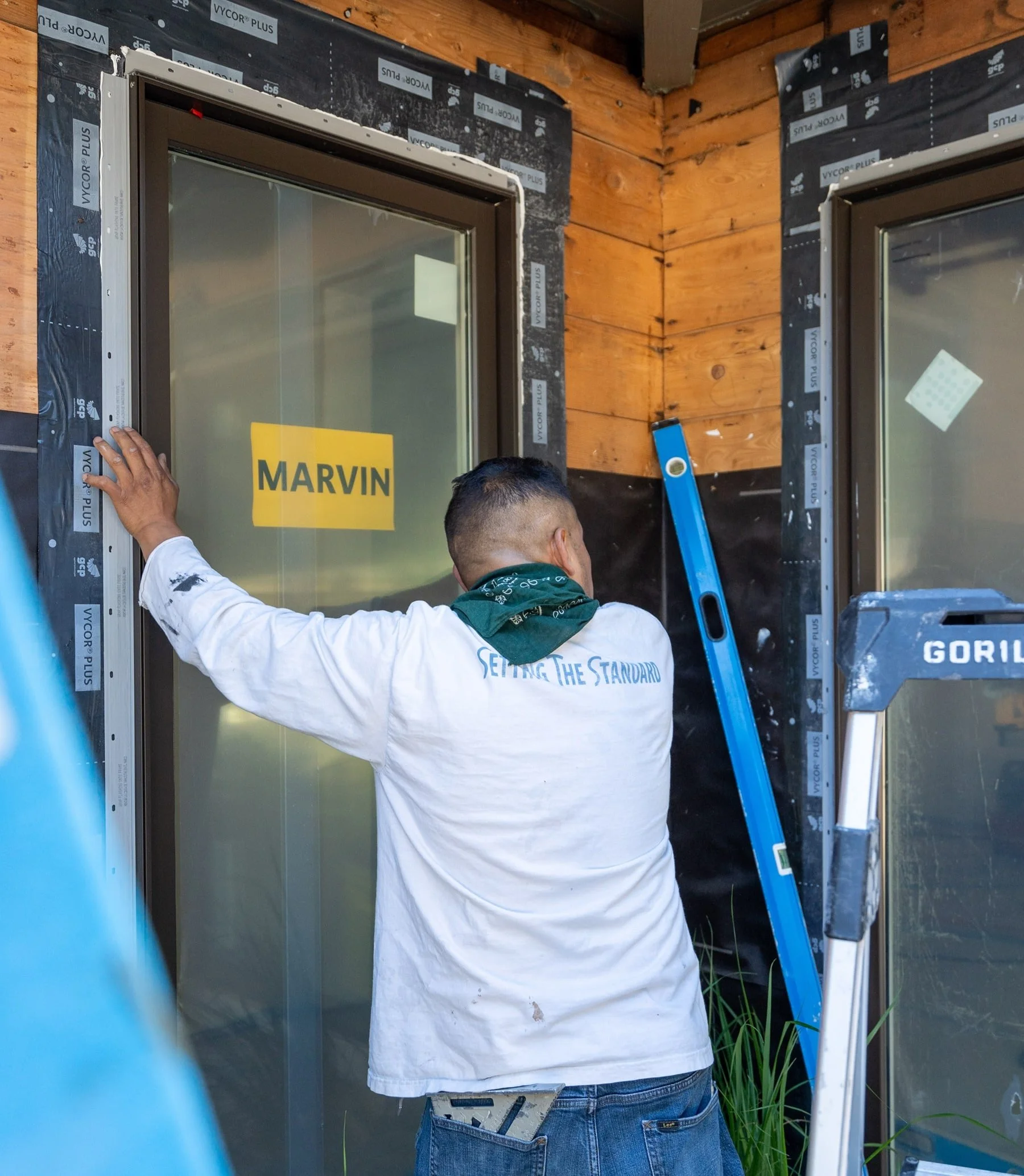A man installing a window with a yellow sign that reads 'MARVIN' on it. The man is wearing a white shirt and has a green bandana around his neck. There are construction materials including a blue level, an aluminum ladder, and black insulation on the wall around him.