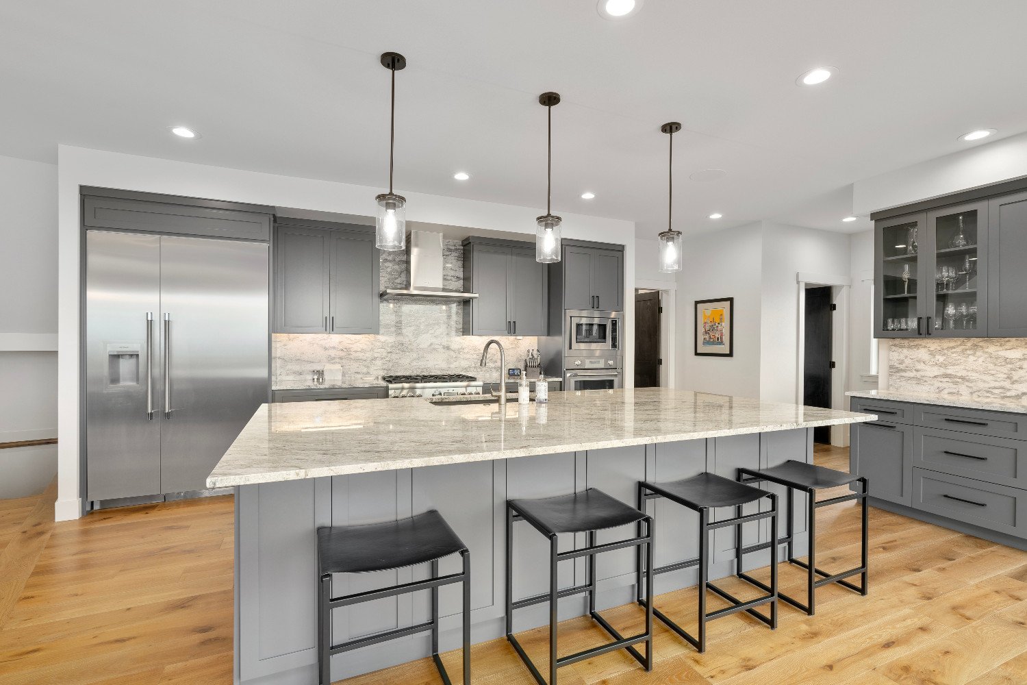 Modern kitchen with gray cabinets, stainless steel appliances, a large island with a granite countertop, black bar stools, and wooden flooring.