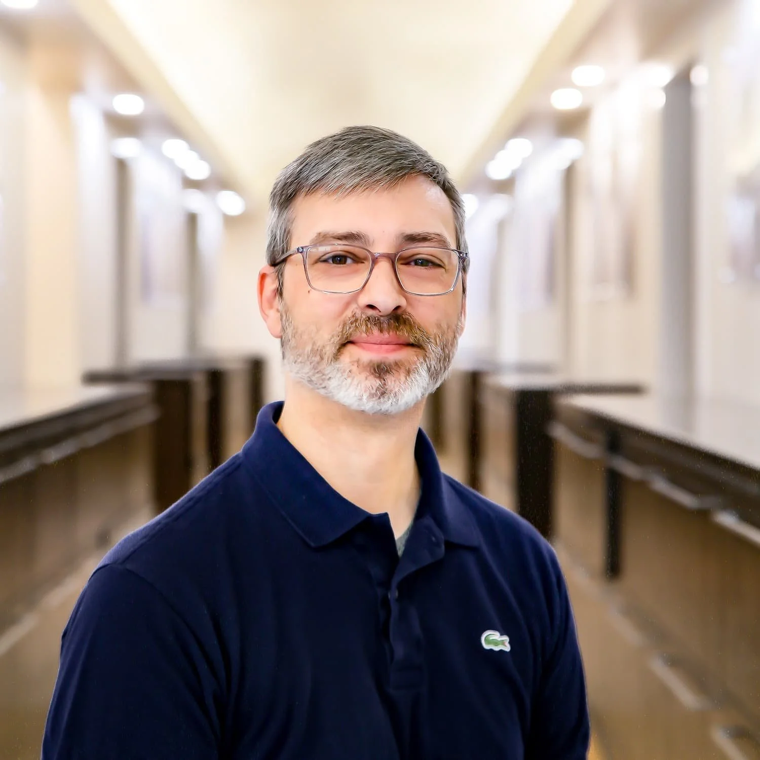 Portrait of a middle-aged man with glasses, short gray hair and beard, wearing a navy blue polo shirt, standing in a well-lit hallway.