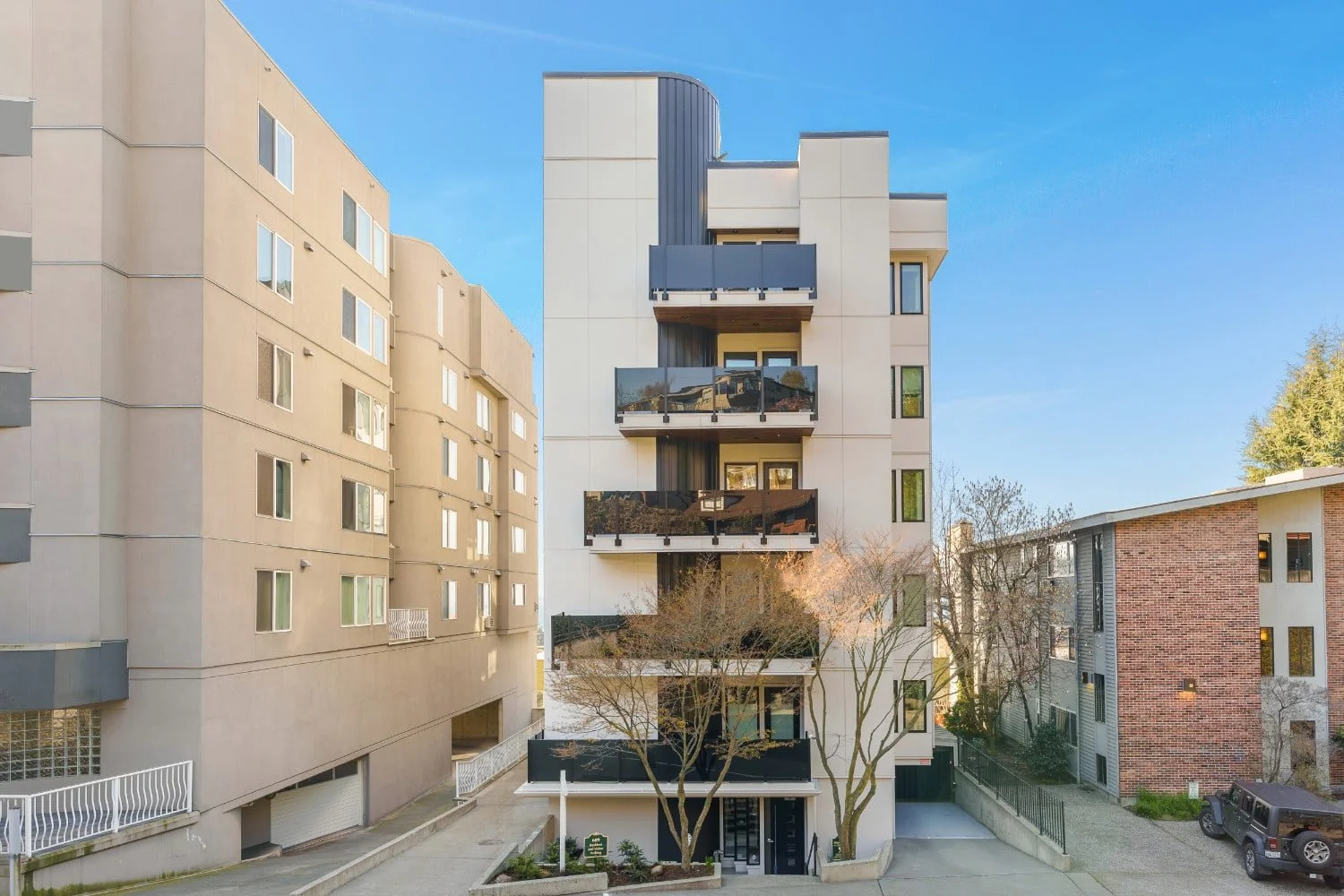 Modern multi-story apartment building with balconies and trees in front, surrounded by other residential buildings, under a clear blue sky.