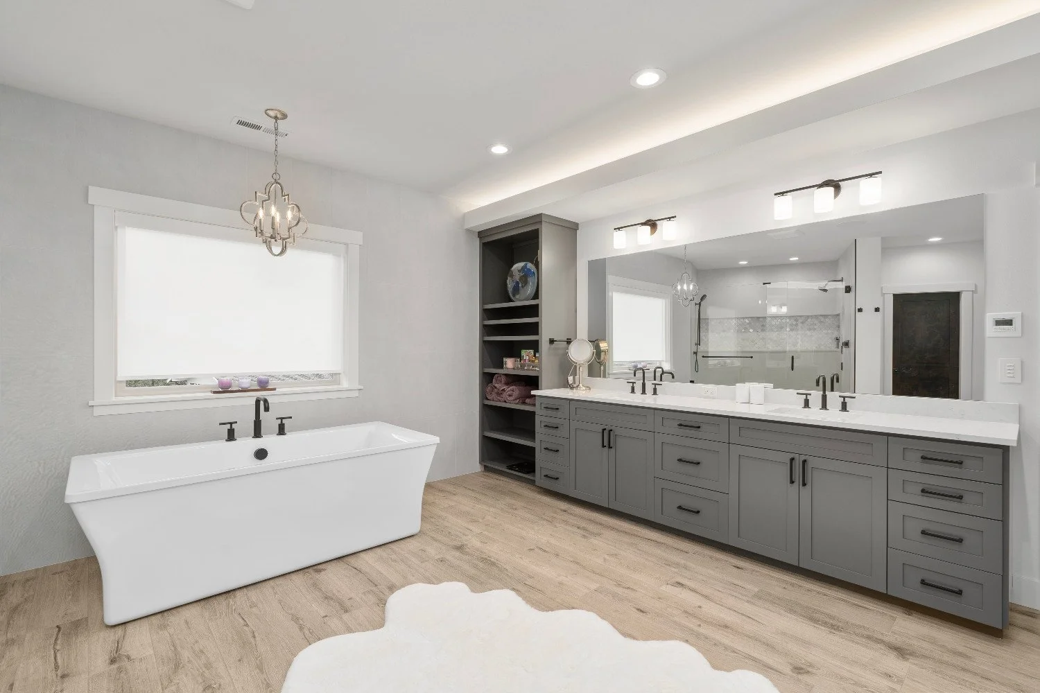 Modern bathroom with a white bathtub under a window, gray cabinetry with black hardware, a large mirror, and a walk-in shower with glass doors.