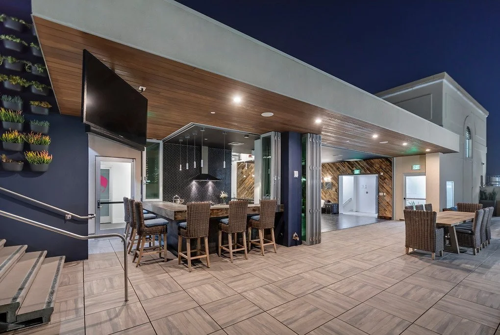 Outdoor rooftop lounge area with high-top seating, a flat-screen TV, and a kitchen bar with pendant lights. There are seating arrangements with chairs and tables, a staircase, potted plants on the wall, and a wood-paneled ceiling.