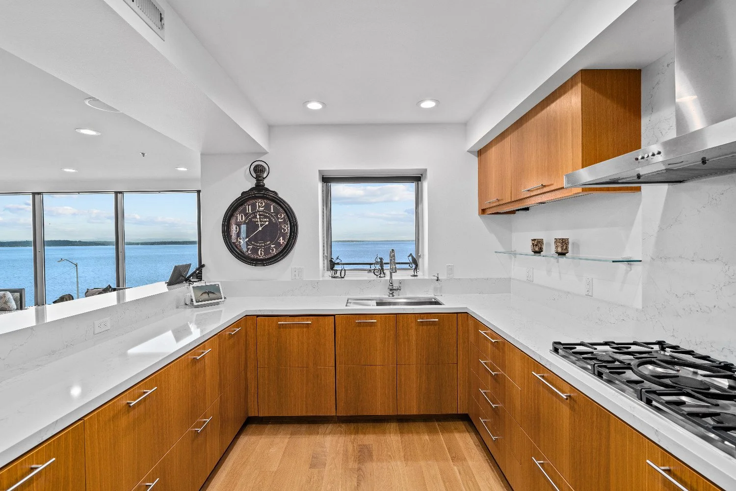 Modern kitchen with wooden cabinets, white marble countertops, a stove, stainless steel vents, and a large clock on the wall. A window shows a view of a body of water and a cloudy sky.