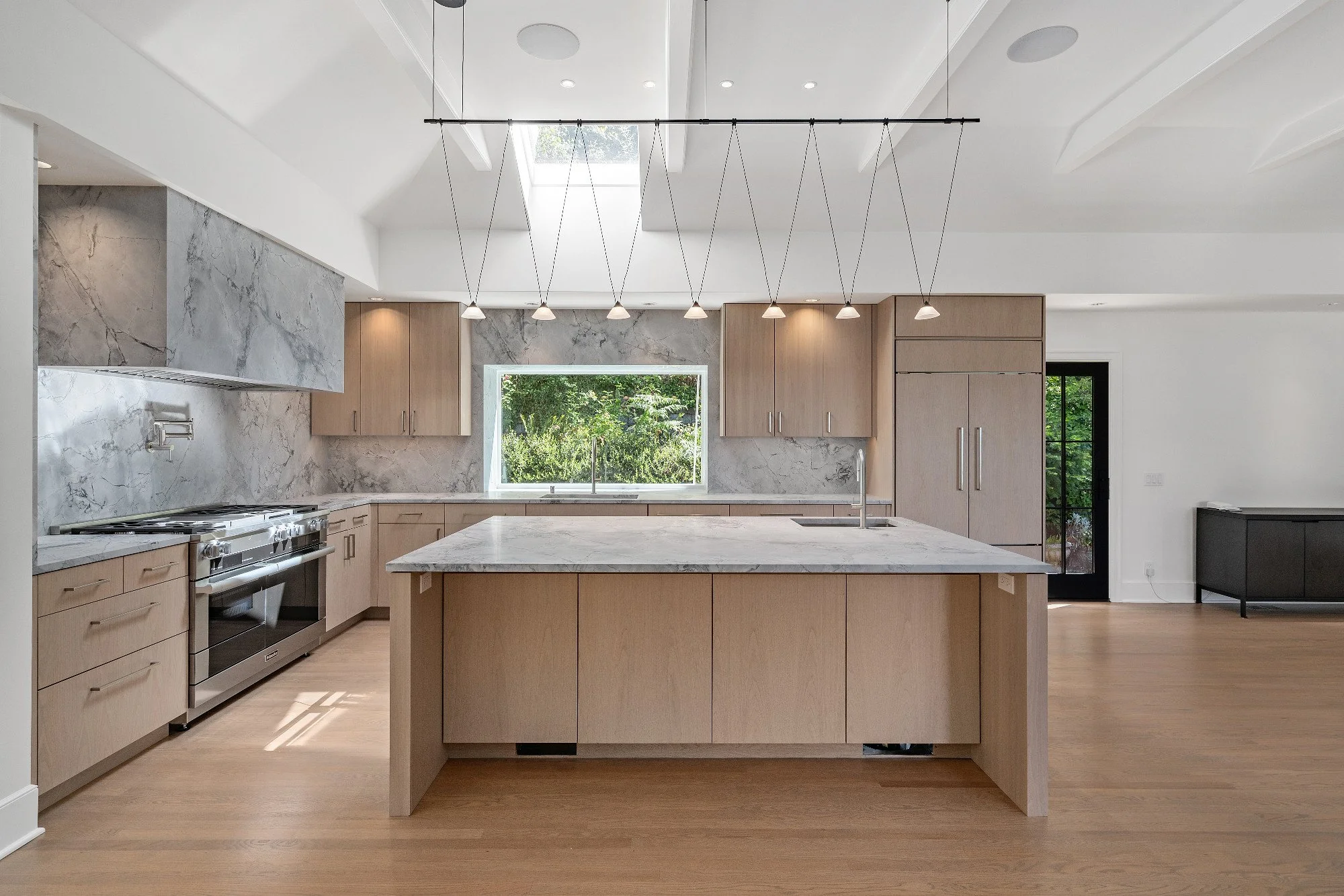 Modern kitchen with marble backsplash and countertops, wooden cabinets, central island, stainless steel stove, and a window showing greenery outside.