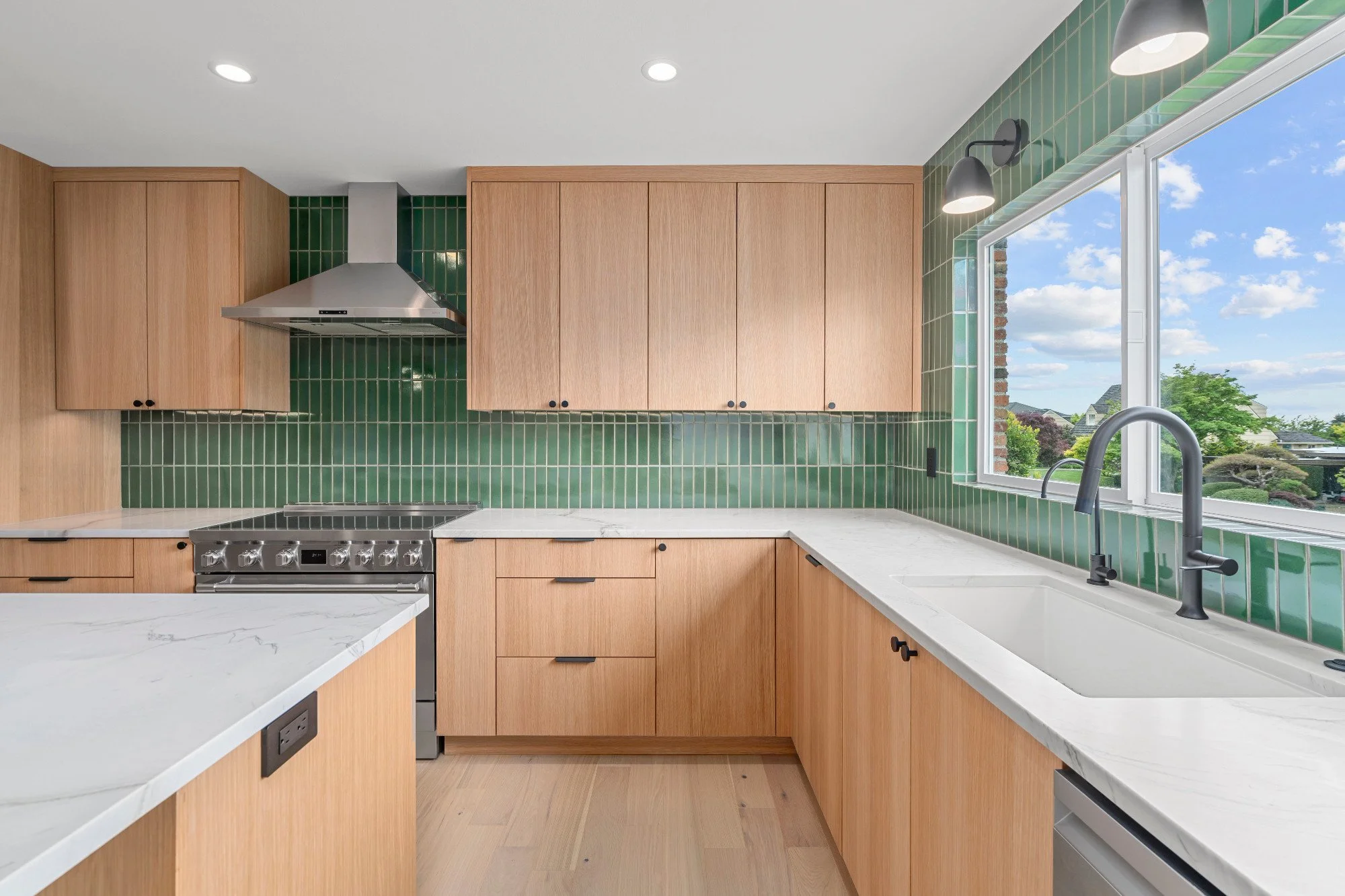 Modern kitchen with wooden cabinets, green tile backsplash, white marble countertops, a stainless steel stove, and a large window overlooking a suburban neighborhood.
