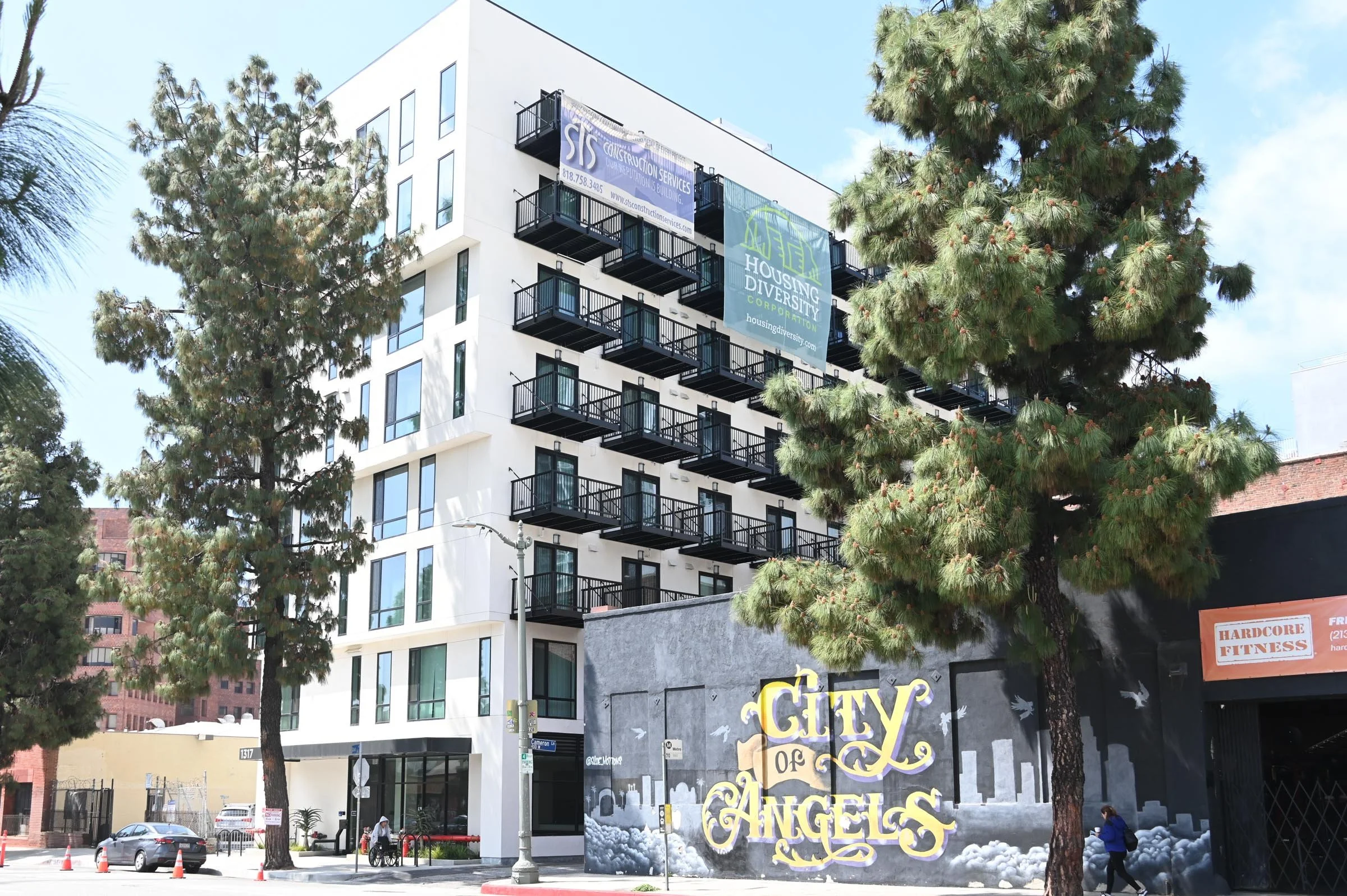 A modern multi-story building with balconies, trees in the foreground, a mural on the wall reading 'City of Angels', and banners advertising construction services and housing diversity.