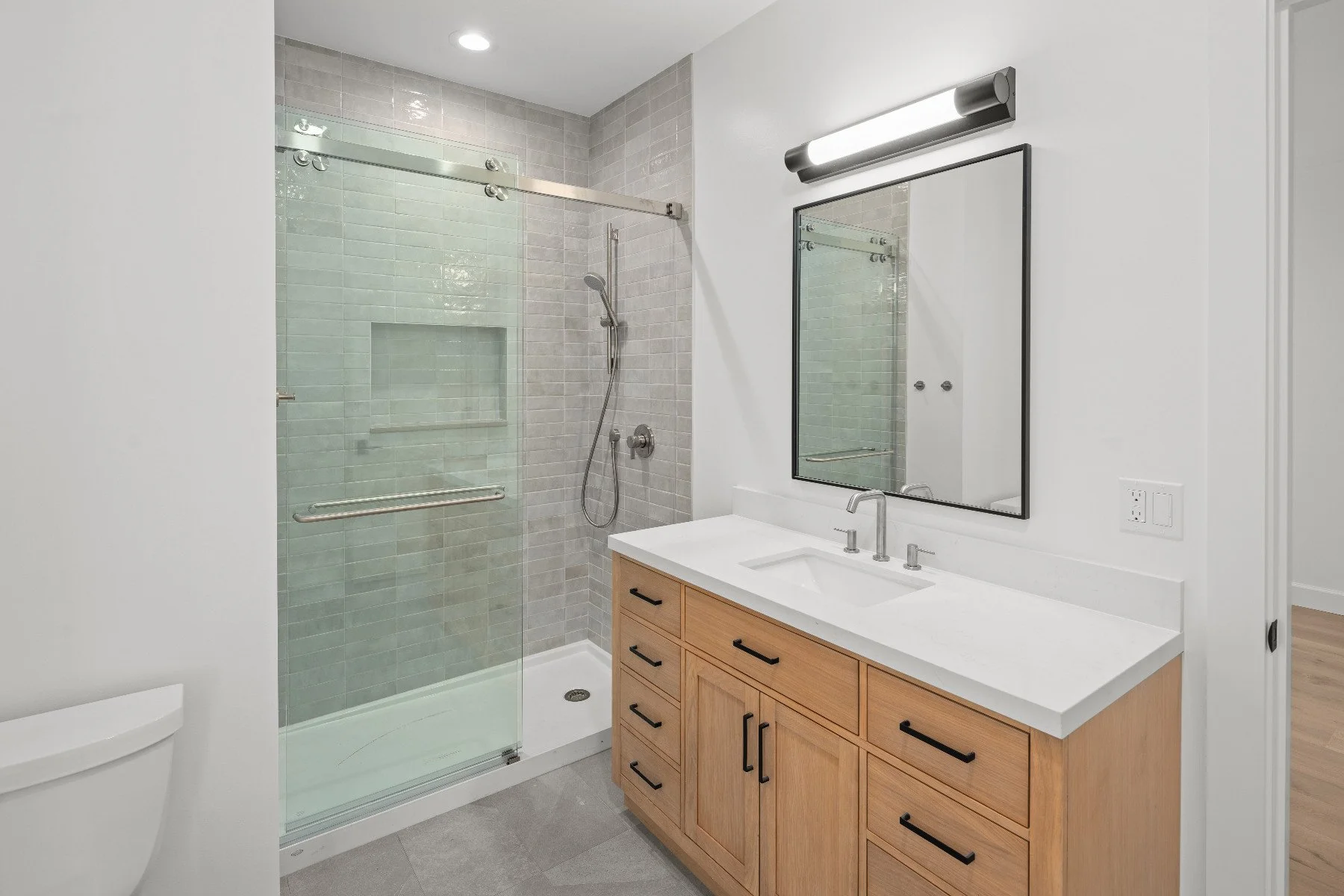 Modern bathroom with a glass-enclosed shower, wooden vanity with a white countertop, a large mirror, and a wall-mounted light fixture.