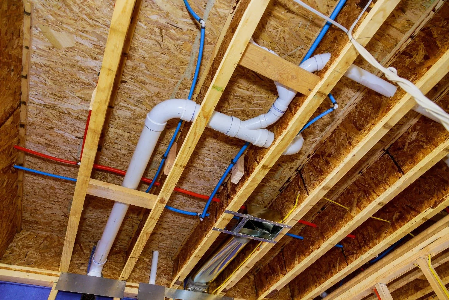 Construction site ceiling showing wooden framing, PVC pipes, electrical conduits, and wiring installation.