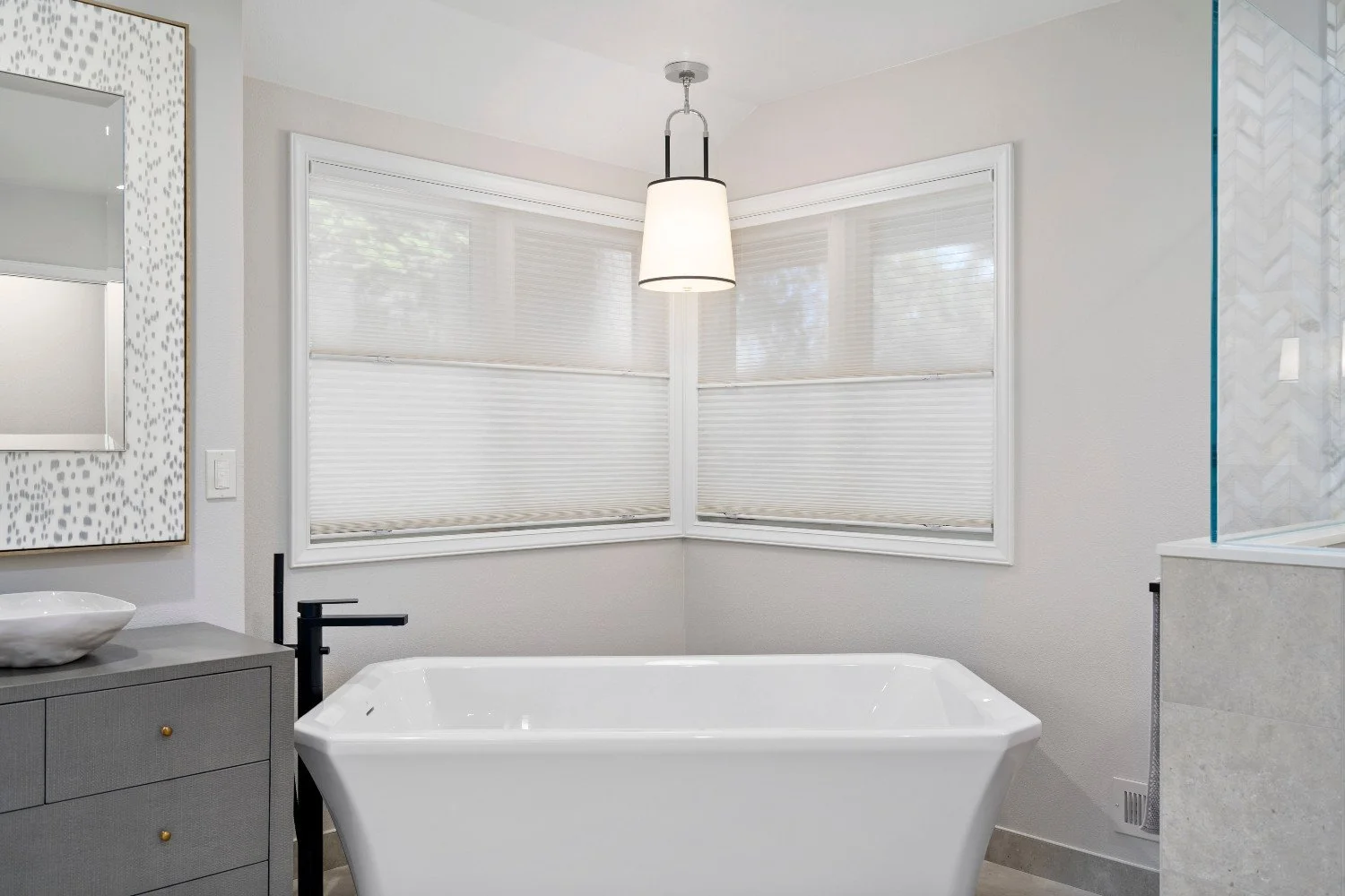 Modern bathroom with a freestanding bathtub, gray vanity with a vessel sink, large window with blinds, pendant ceiling light, and glass shower enclosure.