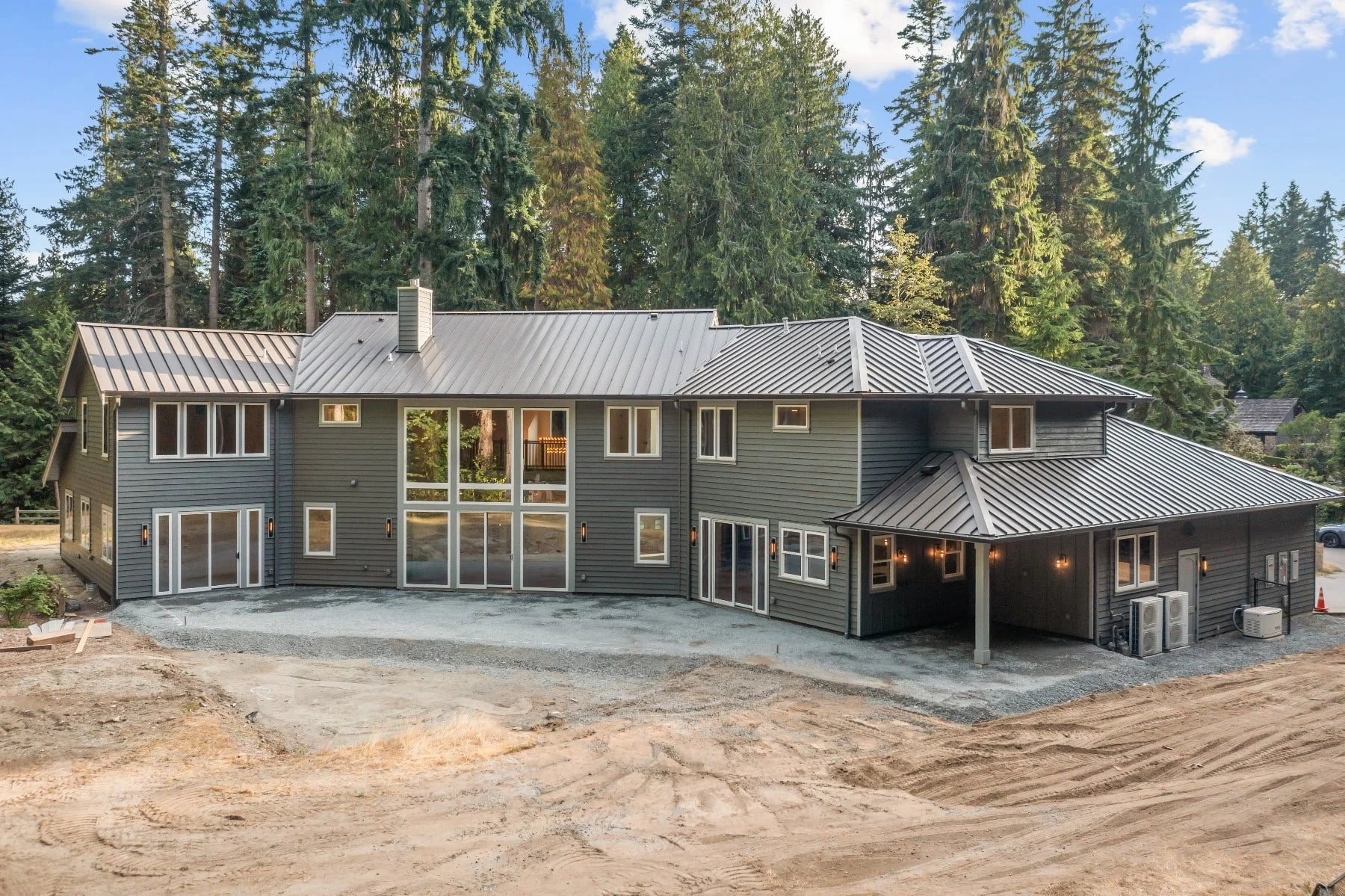A modern two-story house with dark gray siding and a metal roof, surrounded by tall evergreen trees. The house has large windows and sliding glass doors on the ground floor, with some upstairs windows. The area around the house appears to be under co