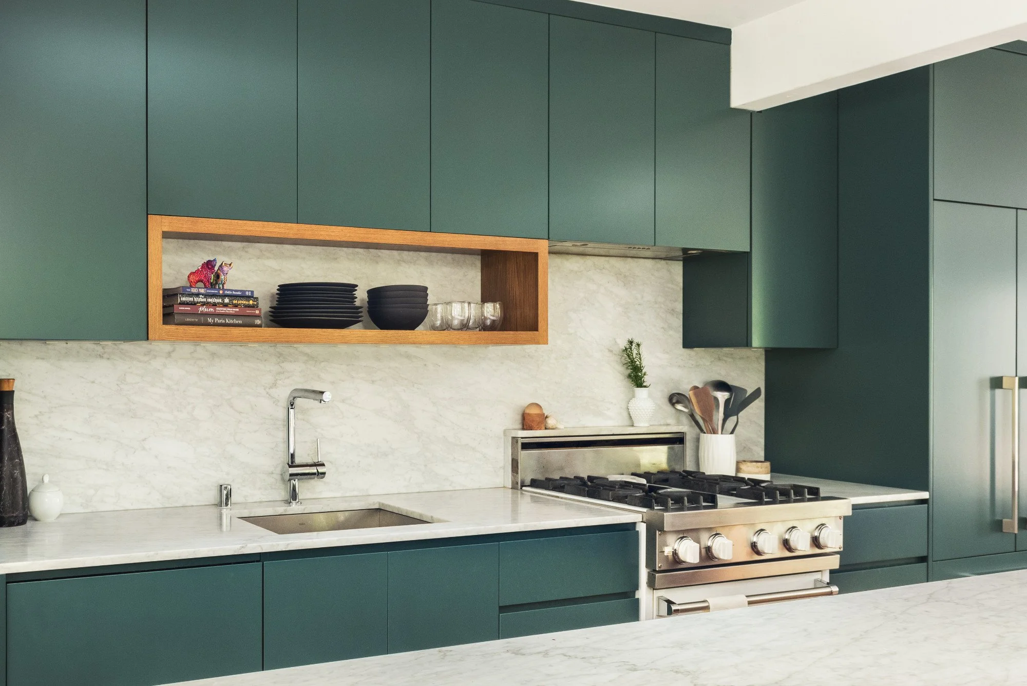 Modern kitchen with green cabinets, white marble countertops and backsplash, stainless steel appliances, open shelf with black dishes and books, small potted plant, and kitchen utensils.