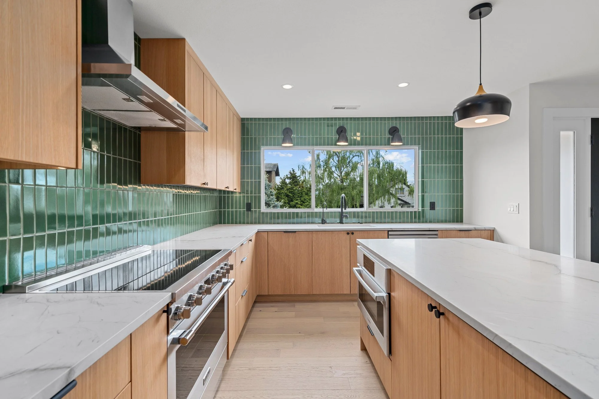 Modern kitchen with green tiled backsplash, wooden cabinets, and white marble countertops. A window above the sink shows trees outside. There are black light fixtures and a black and wood pendant light.