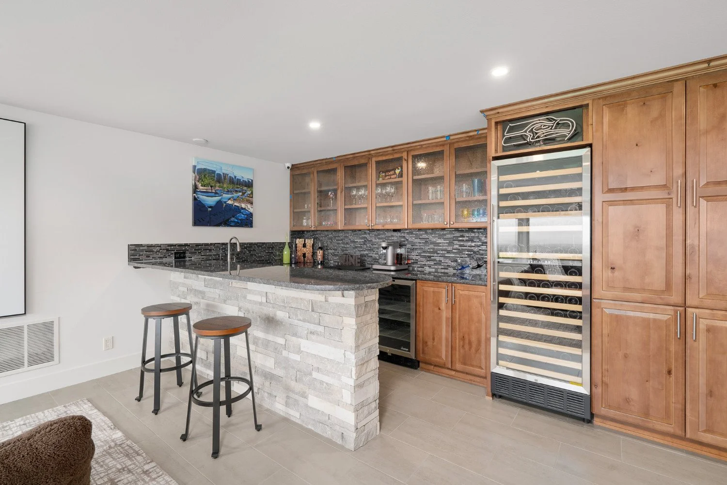 Modern kitchen with a stone island, dark granite countertop, wooden cabinetry, a wine cooler, and a glass-fronted wine rack, with a painting of boats and a city skyline on the wall.