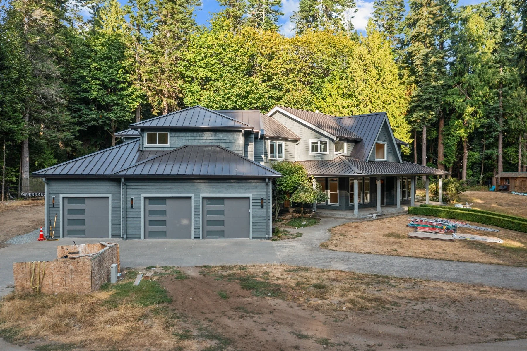 Newly built house with gray siding and metal roof, three car garage, and wooded background.