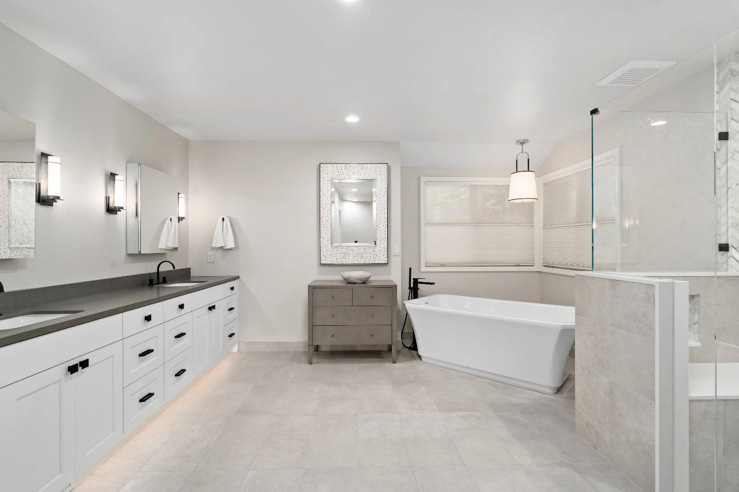 Modern bathroom with white vanity, double sinks, wall-mounted mirrors, towel racks, a freestanding bathtub, a glass-enclosed shower, a dresser with a decorative bowl, and large windows with blinds.