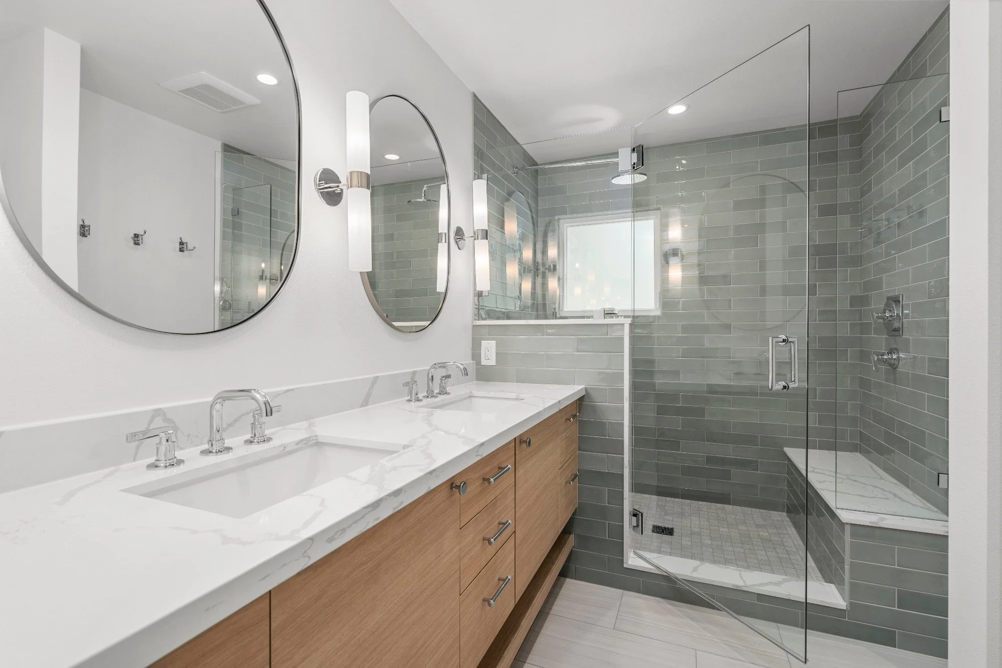 Modern bathroom with double sinks, wooden vanity, oval mirrors, and a glass-enclosed shower with gray tile walls and marble accents.