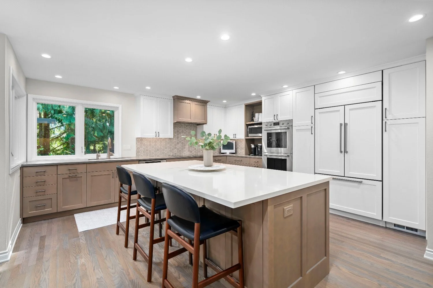 Modern kitchen with white and beige cabinetry, a large kitchen island with a white countertop, black chairs, a window overlooking trees, and built-in appliances.
