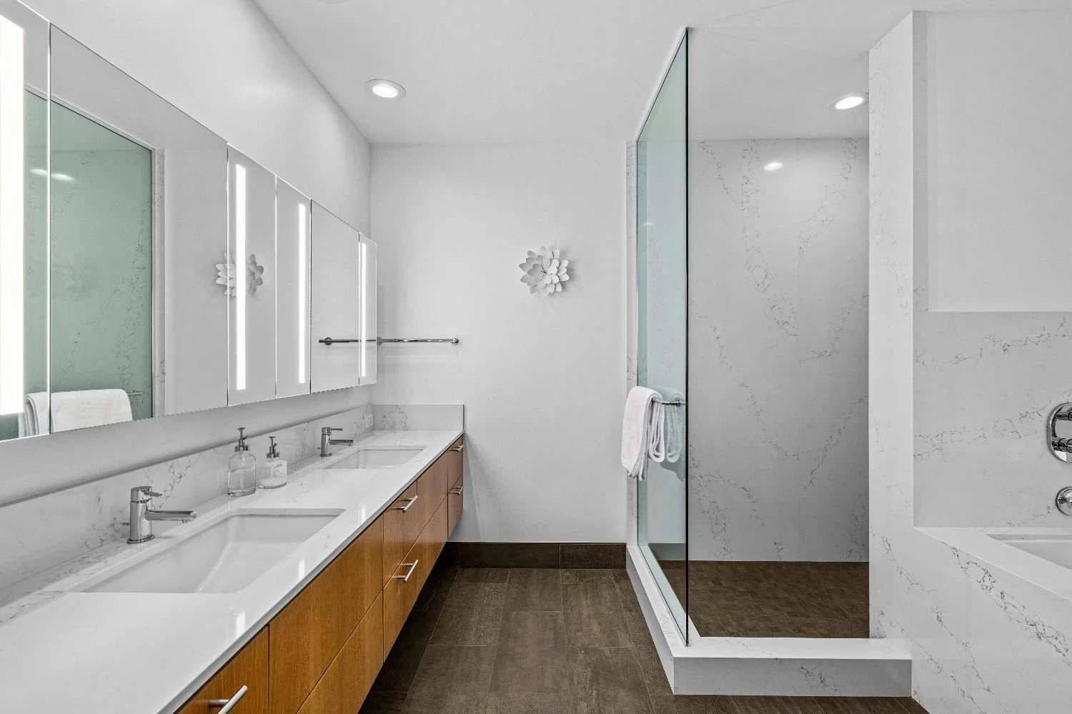 Modern bathroom with double sinks, wood vanity, large mirror with vertical lights, white walls, decorative wall art, glass-enclosed shower, dark floor tiles, and a built-in bathtub.