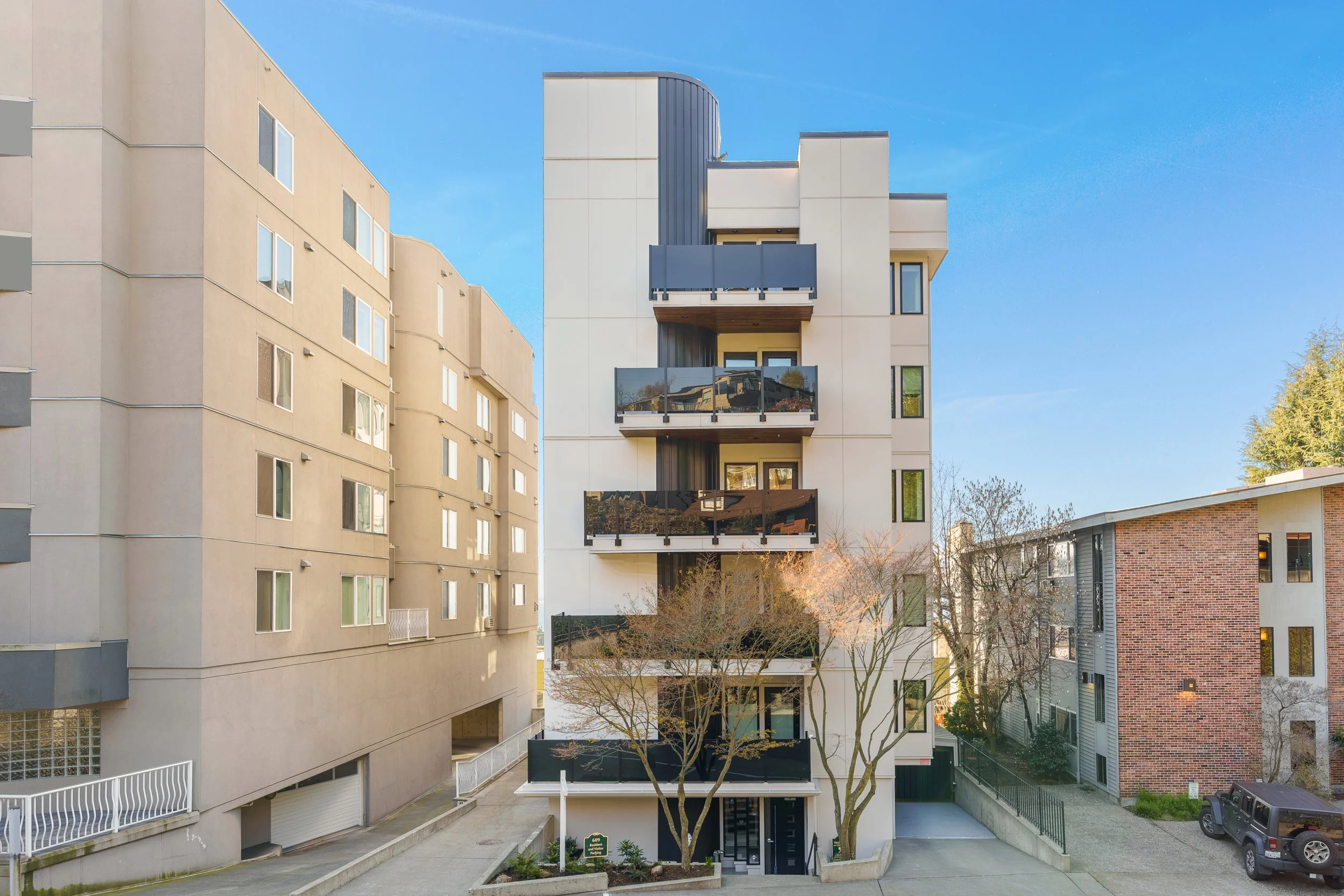 Modern multi-story apartment building with white exterior, black balconies, and large windows, flanked by trees and neighboring buildings, under a clear blue sky.