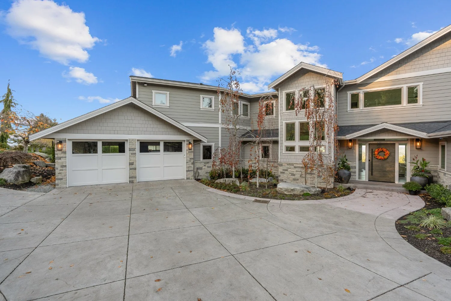 A modern two-story house with a driveway, a two-car garage, and a front porch decorated with a fall wreath. The house has light gray siding, stone accents, and large windows. There are small trees and potted plants near the entrance, under a partly c