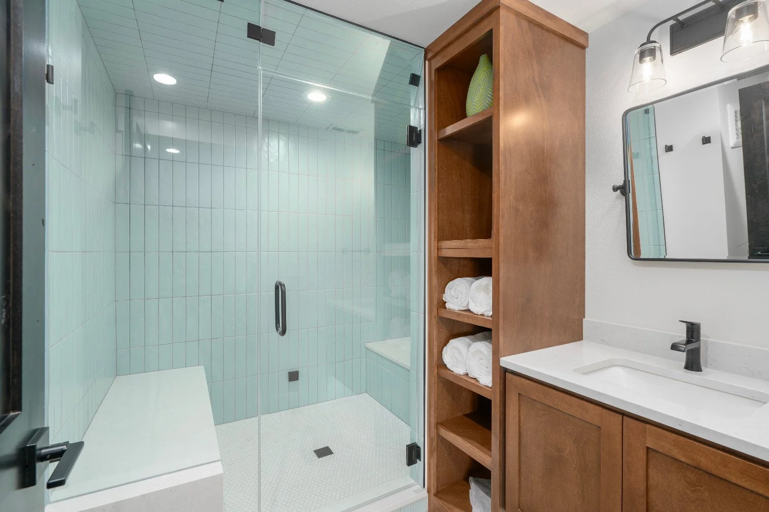 Modern bathroom with a glass-enclosed shower, wooden cabinet with open shelving, a white countertop with a sink, and a mirror.