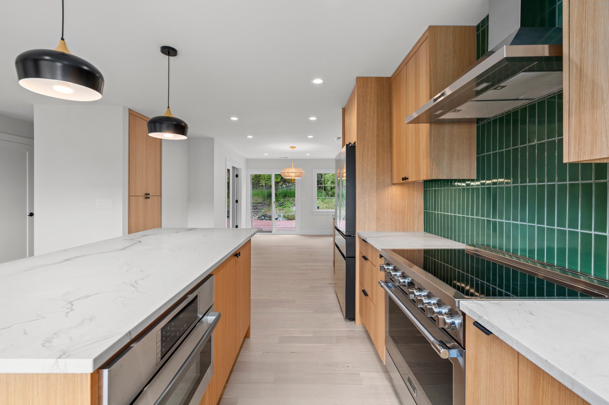 Modern kitchen with white marble countertops, black pendant lights, green tiled backsplash, wooden cabinets, and stainless steel appliances.