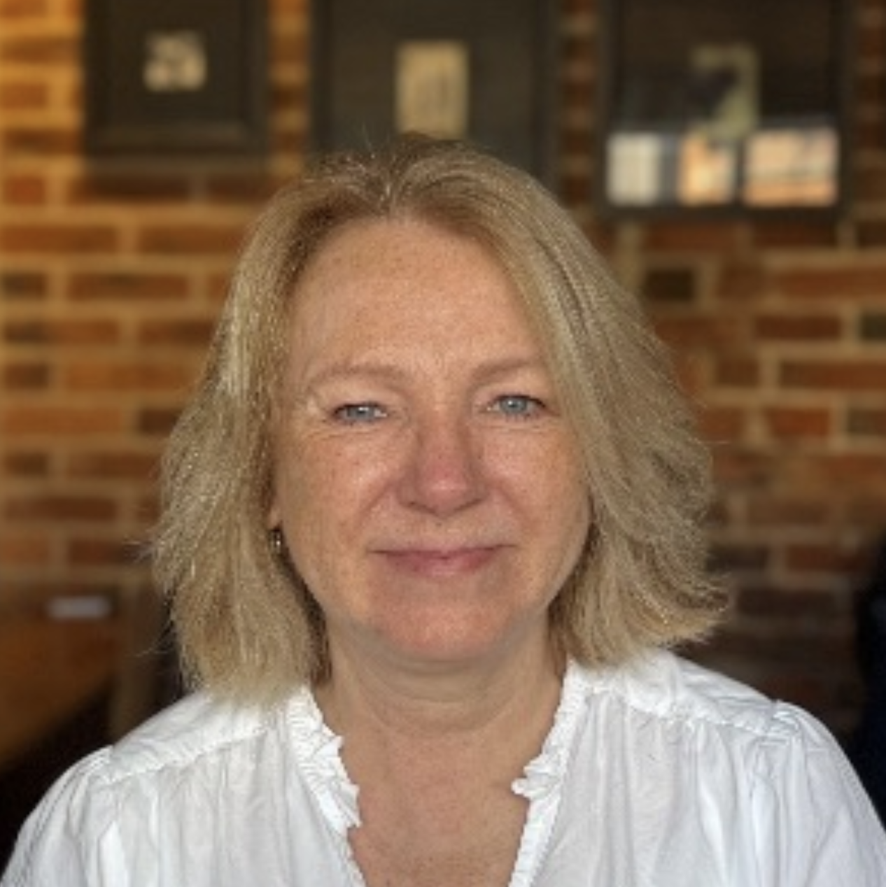 A smiling woman with blonde hair and blue eyes, wearing a white blouse, sitting in a room with a brick wall background and framed pictures.