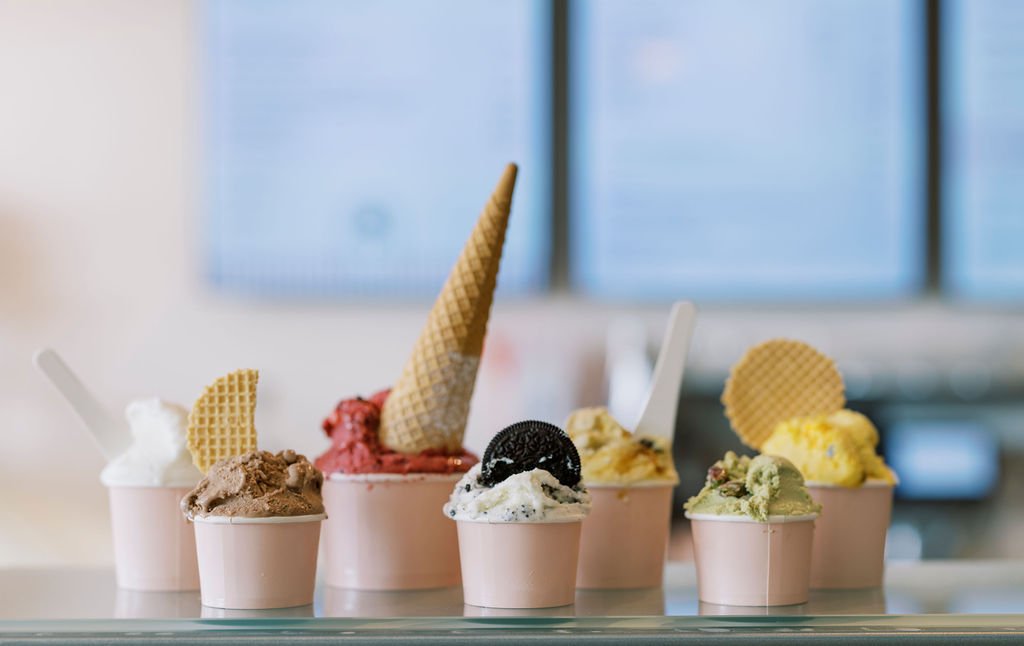 Six cups of various ice cream flavors with toppings and waffle cones on a counter in a cafe.