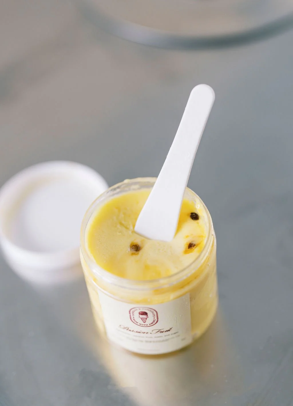 A jar of lemon-flavored ice cream or gelato with black peppercorns, a white spatula, and a white lid in the background, sitting on a gray surface.