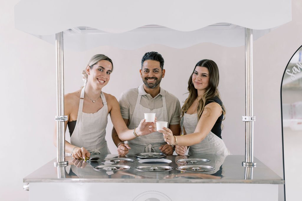 Three people in aprons serving and enjoying ice cream at a small ice cream stand in a bright, modern setting.