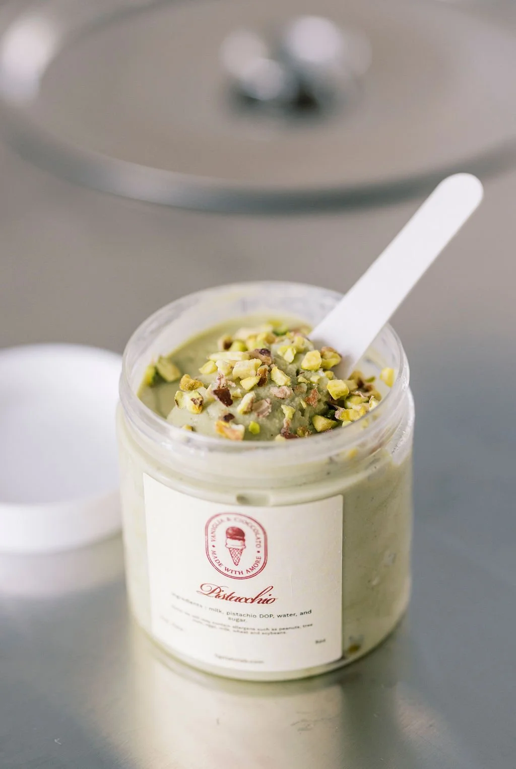 A jar of pistachio gelato topped with chopped pistachios and a white spoon, placed on a gray surface with a blurred background.