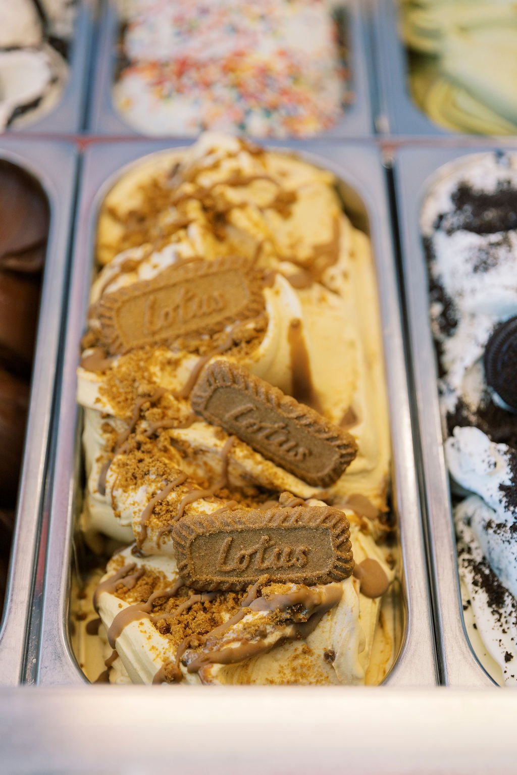 A close-up of a tub of vanilla ice cream topped with graham cracker pieces, chocolate drizzle, and Oreo cookie halves, in an ice cream shop freezer.