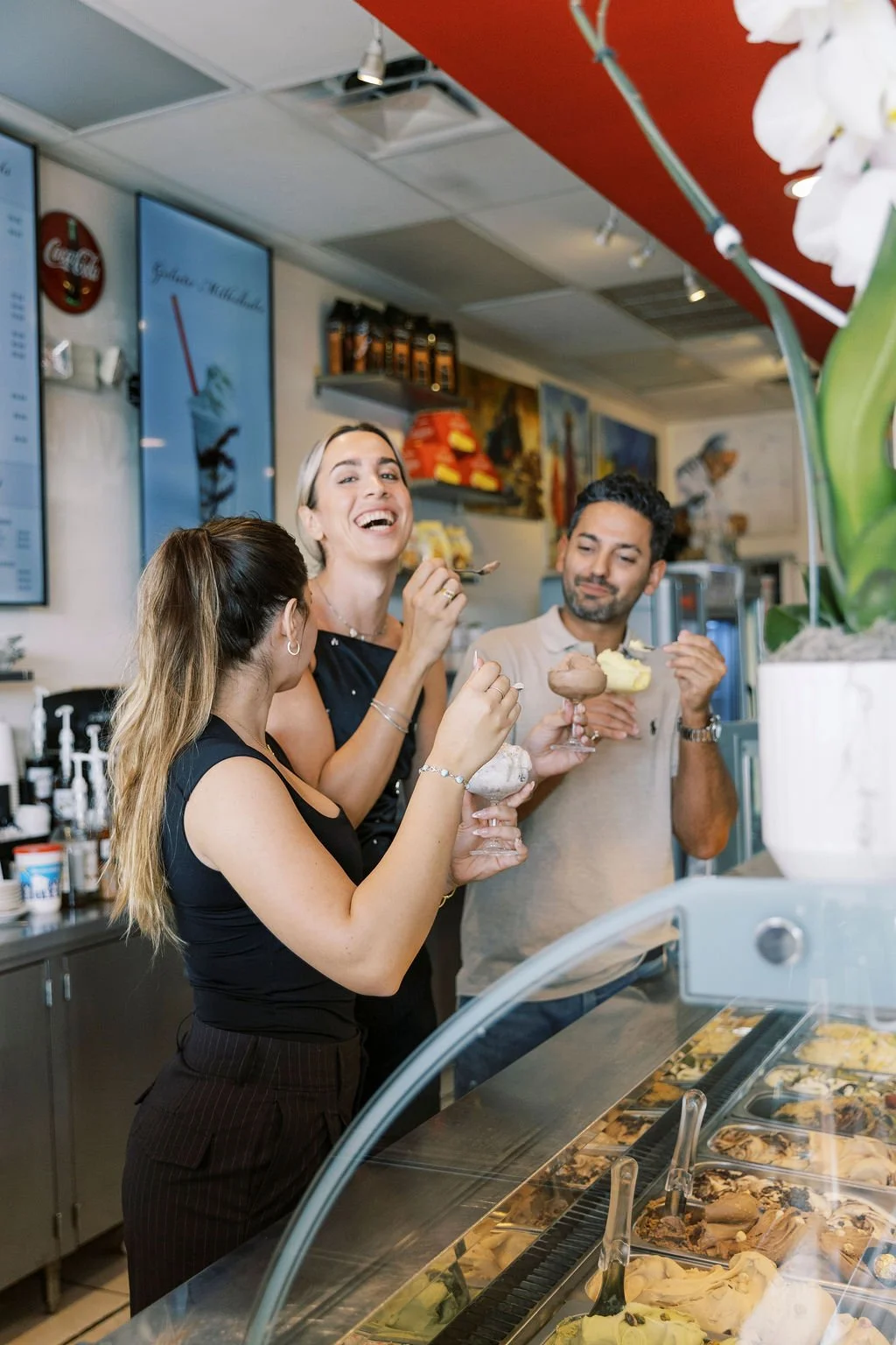 Three people happily enjoy ice cream inside an ice cream shop, with a display case of various ice cream flavors in front of them.