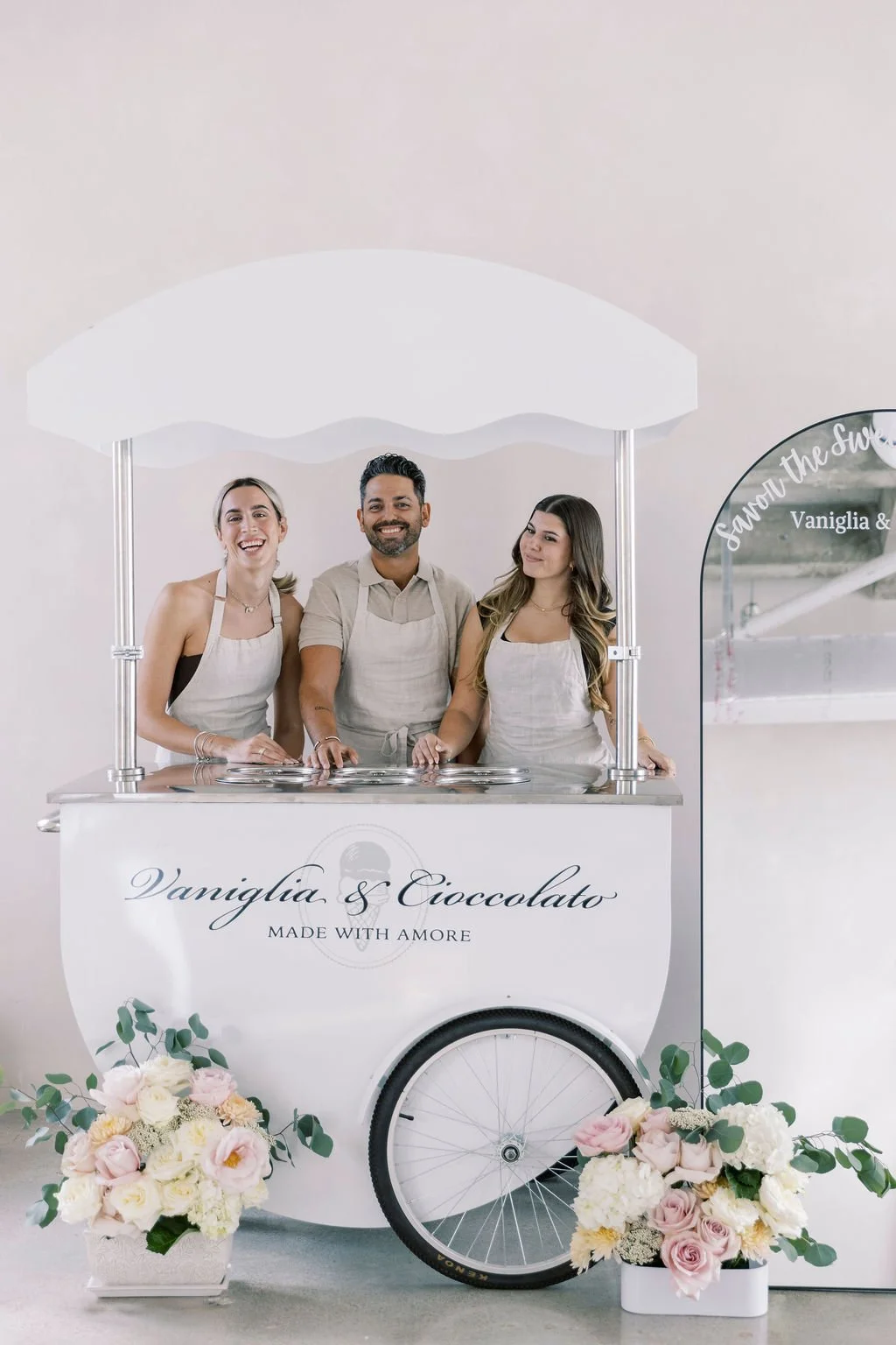 Three smiling people in aprons standing behind a white ice cream cart decorated with flower arrangements, inside a light-colored indoor space.